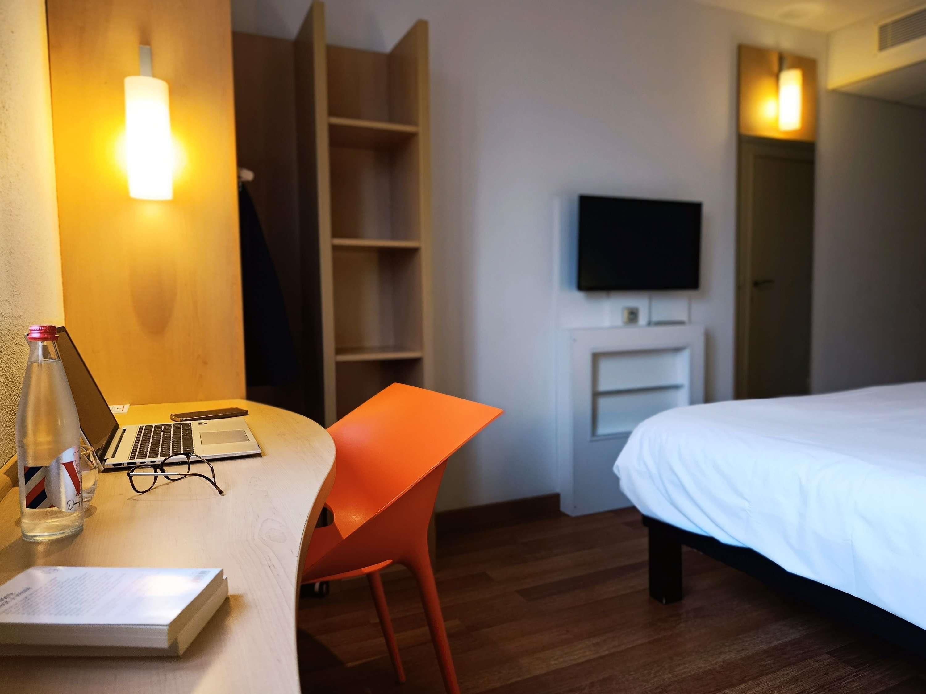 Una camera d'albergo con un letto, una scrivania con un portatile, una sedia, un televisore montato a parete e un armadio.