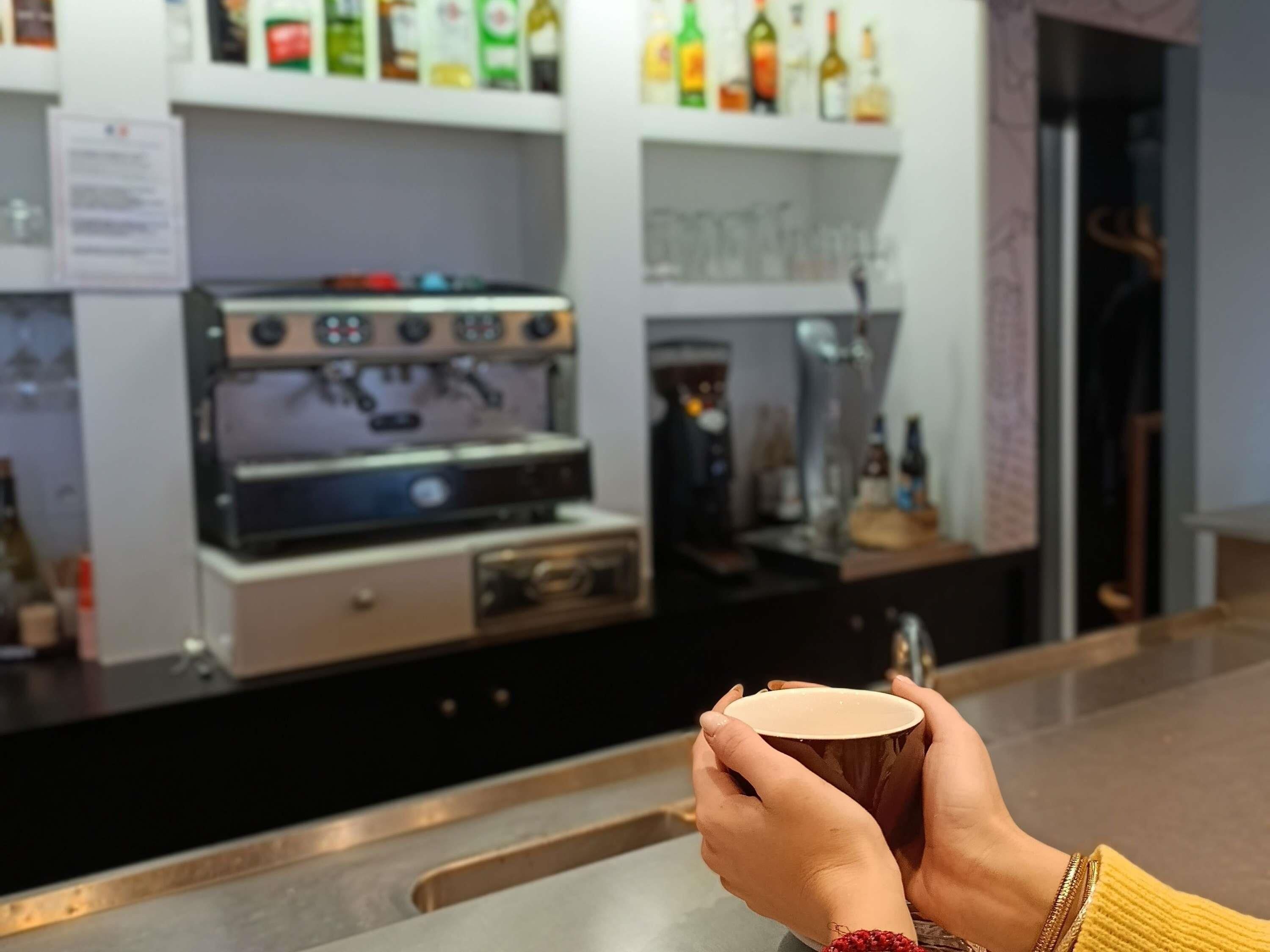 Un barista sostiene una taza, con máquinas de espresso y estantes llenos de bebidas al fondo.