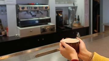 Un barista sostiene una taza, con máquinas de espresso y estantes llenos de bebidas al fondo.