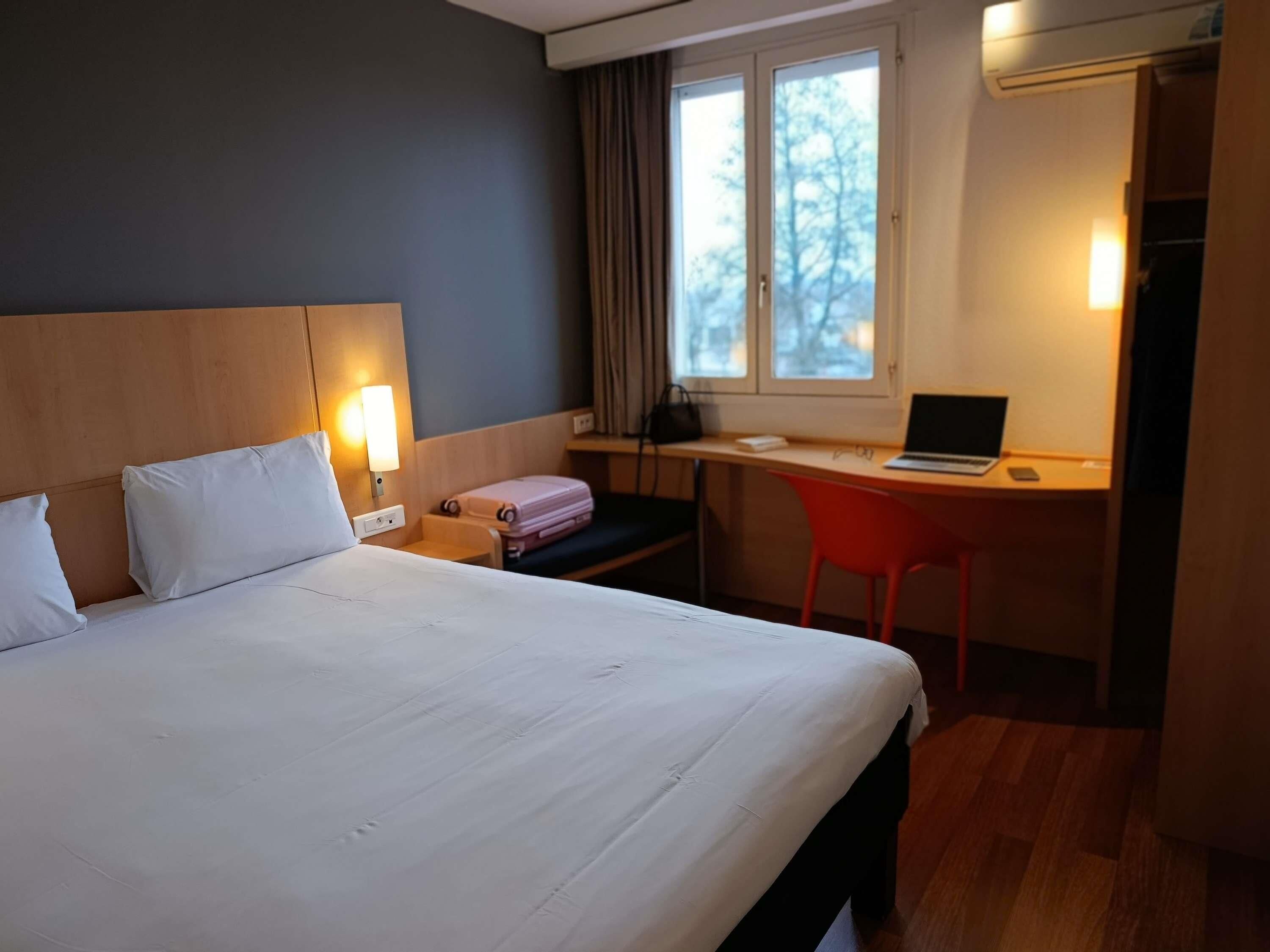 A hotel room with a bed, a desk with a laptop, a chair, a suitcase, and a window with curtains.