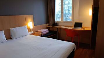 A hotel room with a bed, a desk with a laptop, a chair, a suitcase, and a window with curtains.