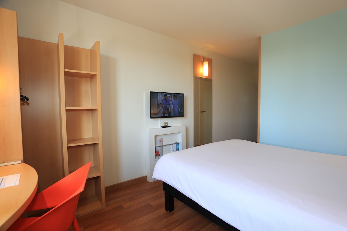 A hotel room with a bed, a desk, a chair, a TV, and a wardrobe.