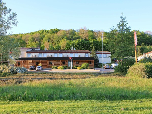 A two-story building with a flat roof, a visible entrance, and a parking area with cars. - ibis Aurillac (Aurillac)
