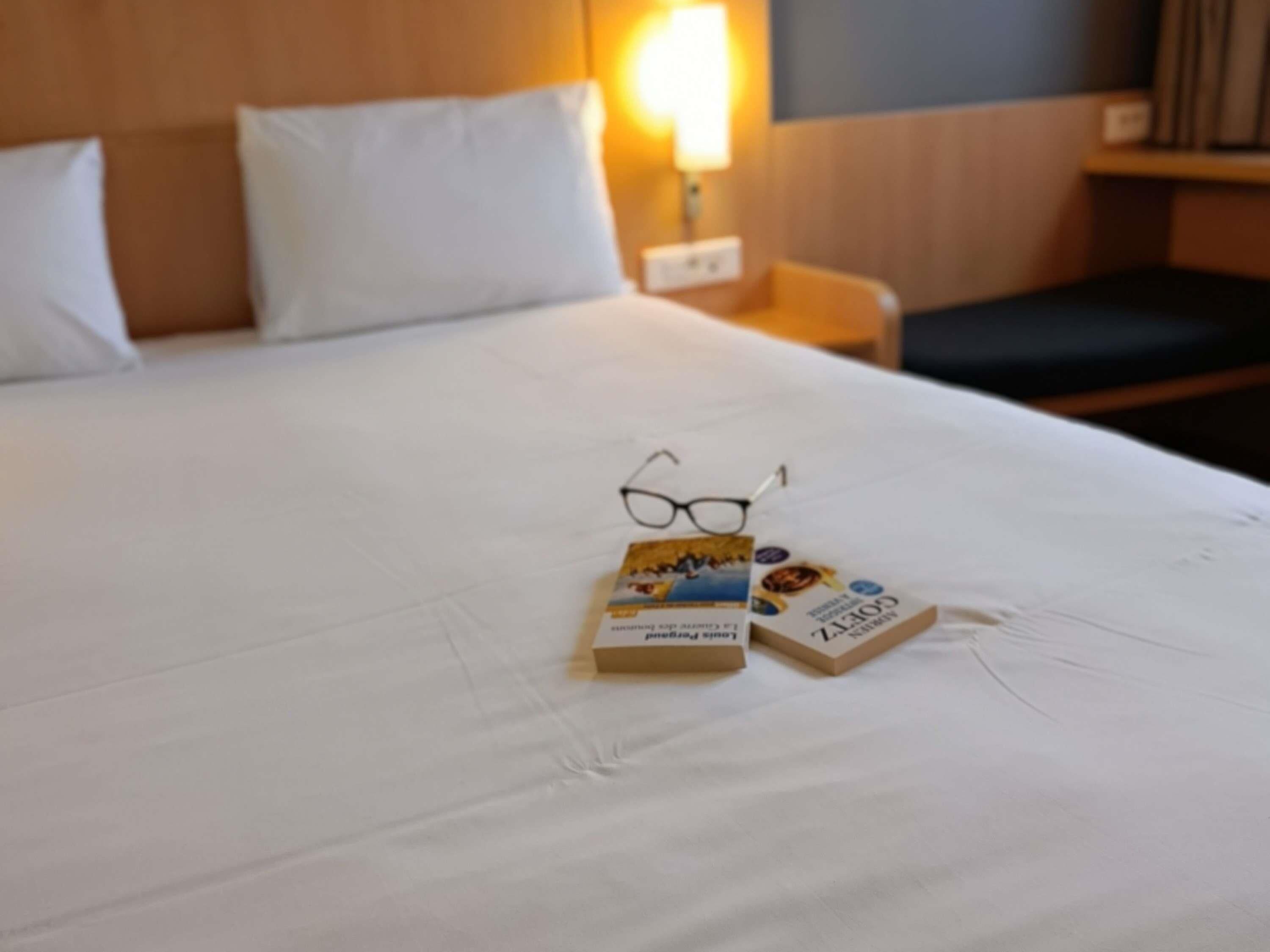 A neatly made bed with white linens, a pair of glasses, and two books on the bed.