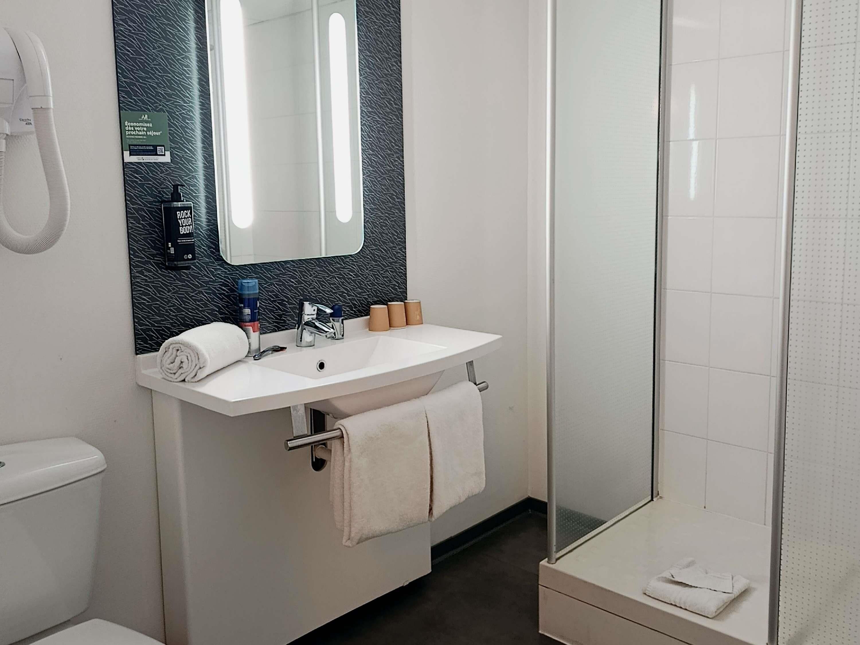 Un baño con lavamanos, espejo y inodoro blancos. Se observa un perchero con toallas, una zona de ducha y una botella de champú Rock Your Body!