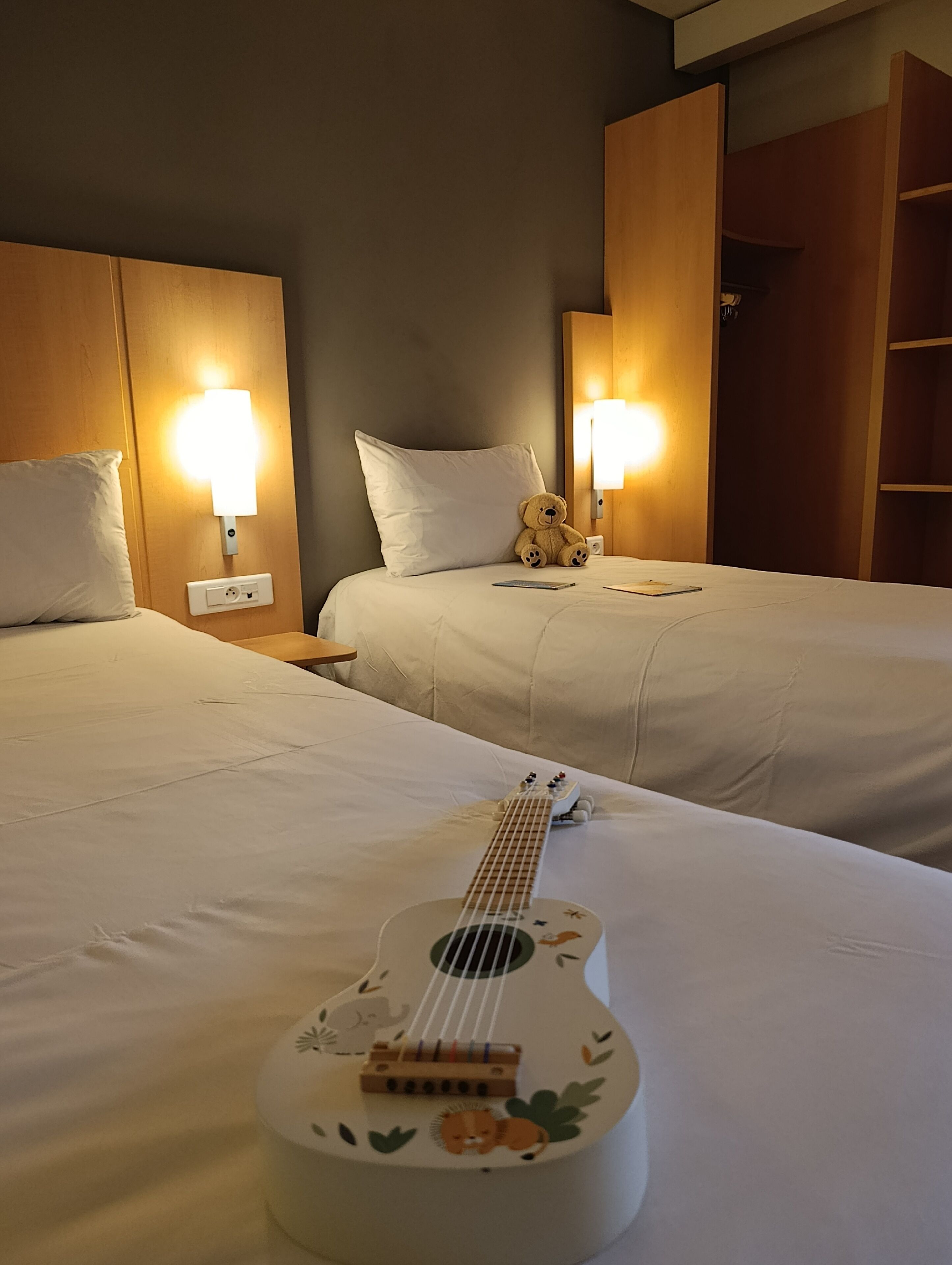 Habitación de hotel con dos camas, una guitarra sobre la cama y un osito de peluche en el cabecero.