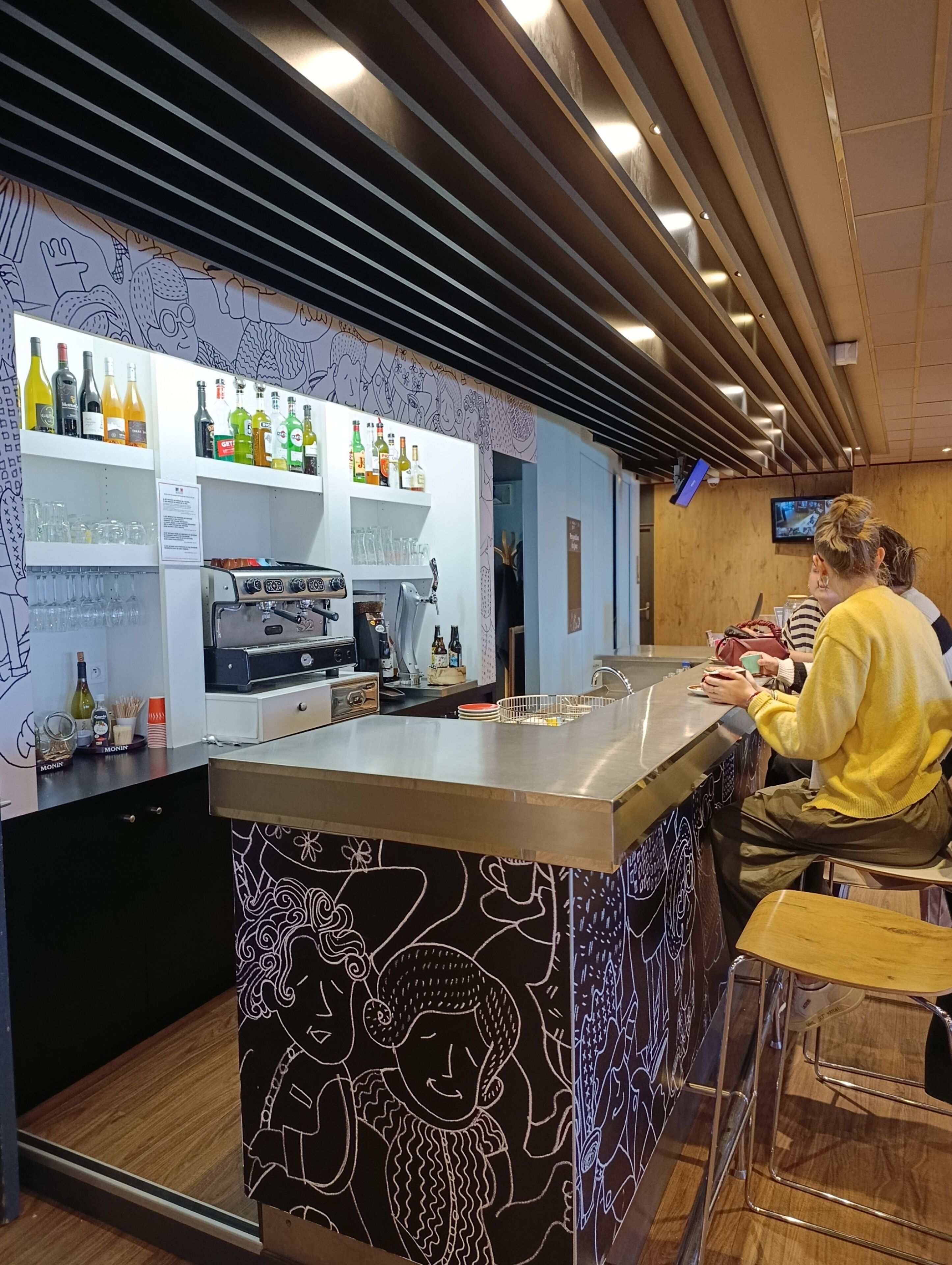 Una zona de bar con mostrador, estanterías con botellas, vasos y una pantalla de televisión.