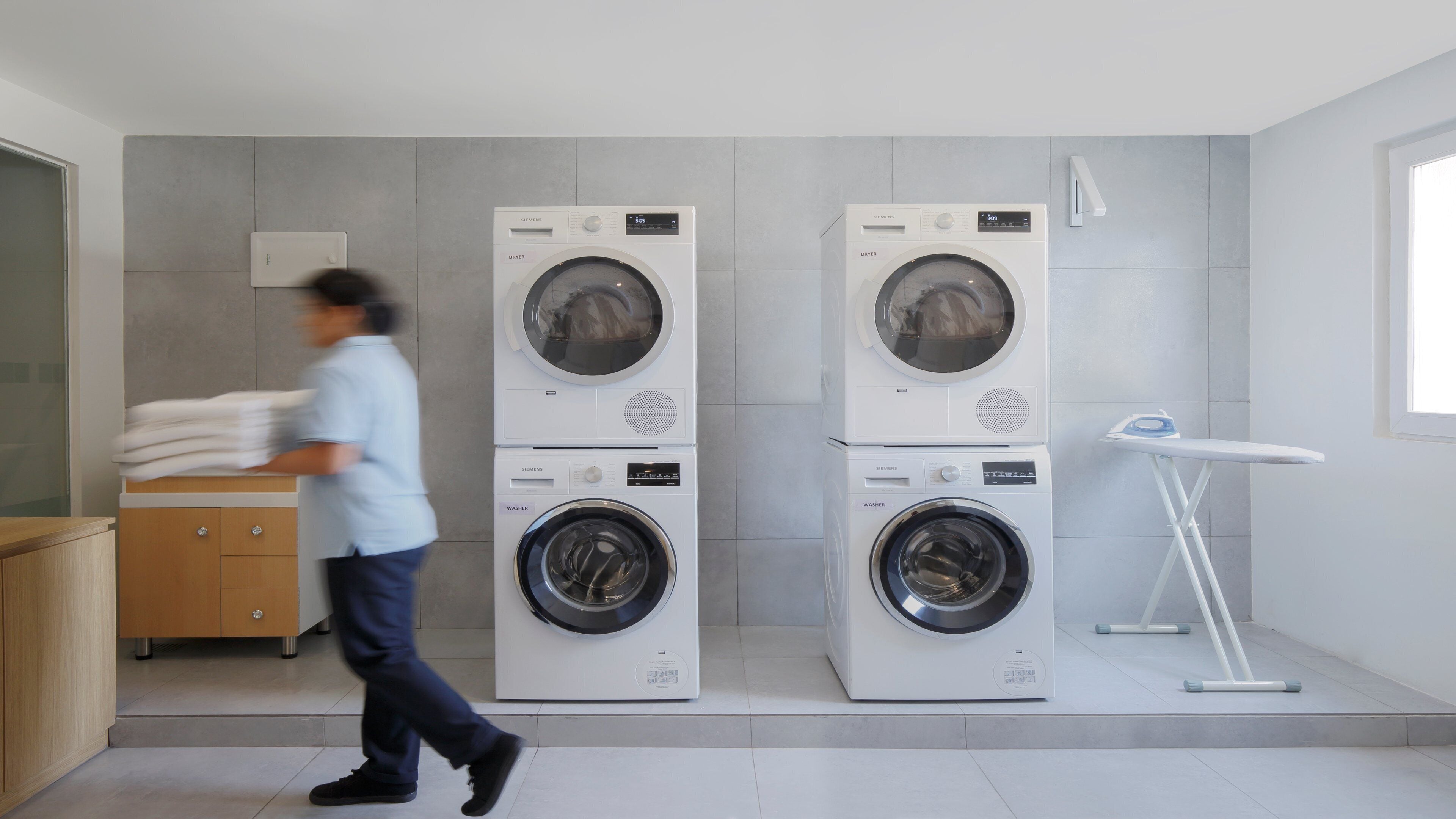 A modern laundry room with two stacked washer-dryer units, a wooden cabinet, and a small ironing station.