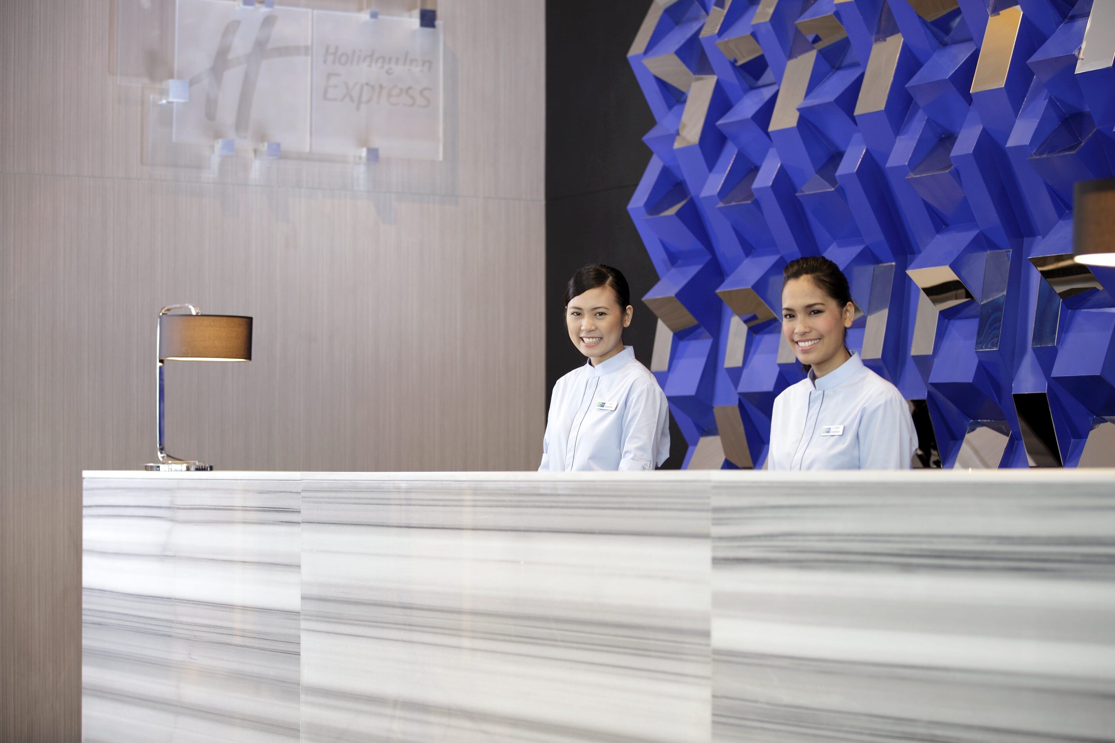 A reception desk with a modern design, a sign indicating 'Holiday Inn Express', and a geometric blue wall feature.