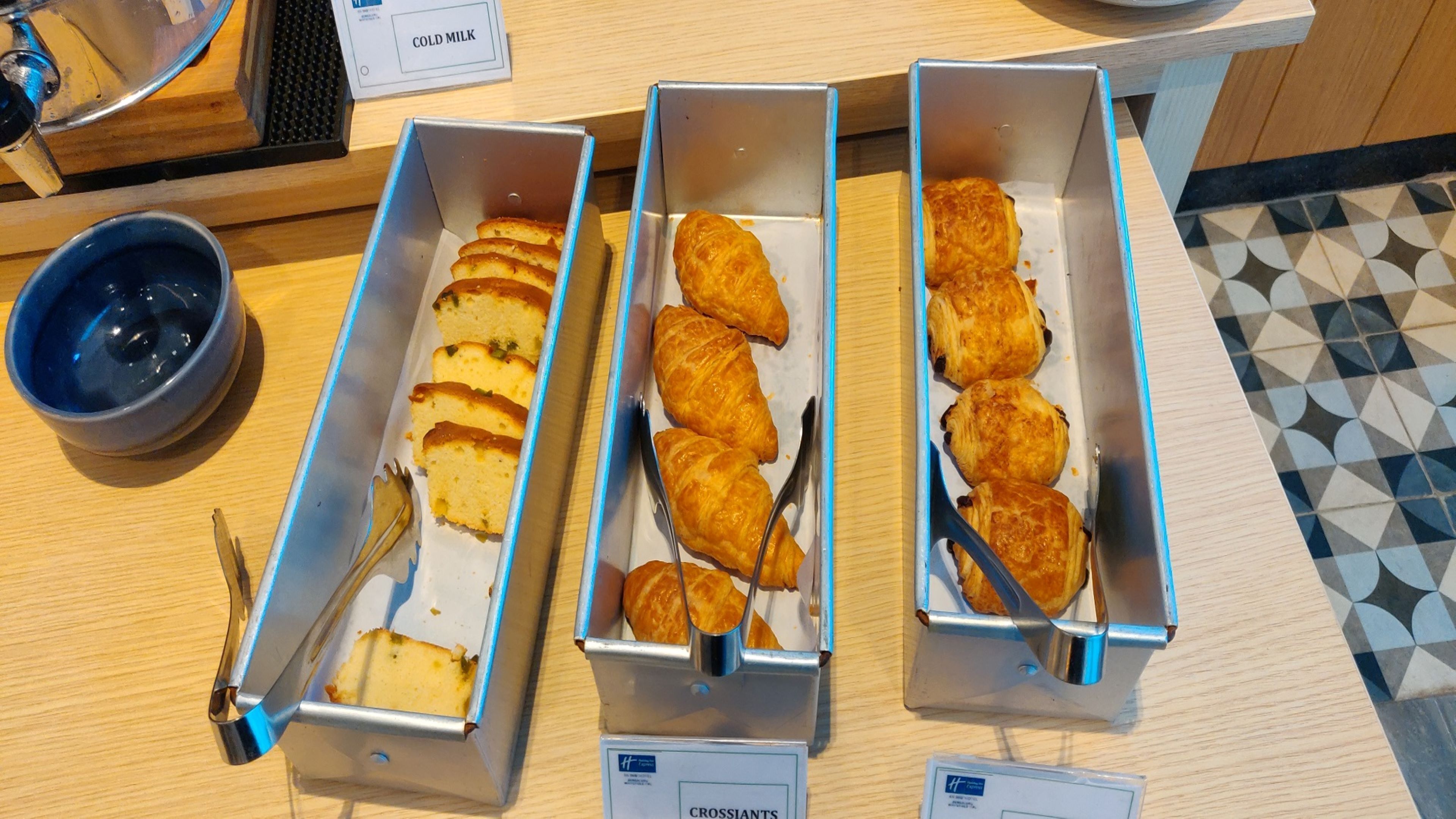 Trois plateaux en métal contenant des aliments, une pancarte indiquant « LAIT FRAIS » et un bol bleu sur un comptoir en bois.