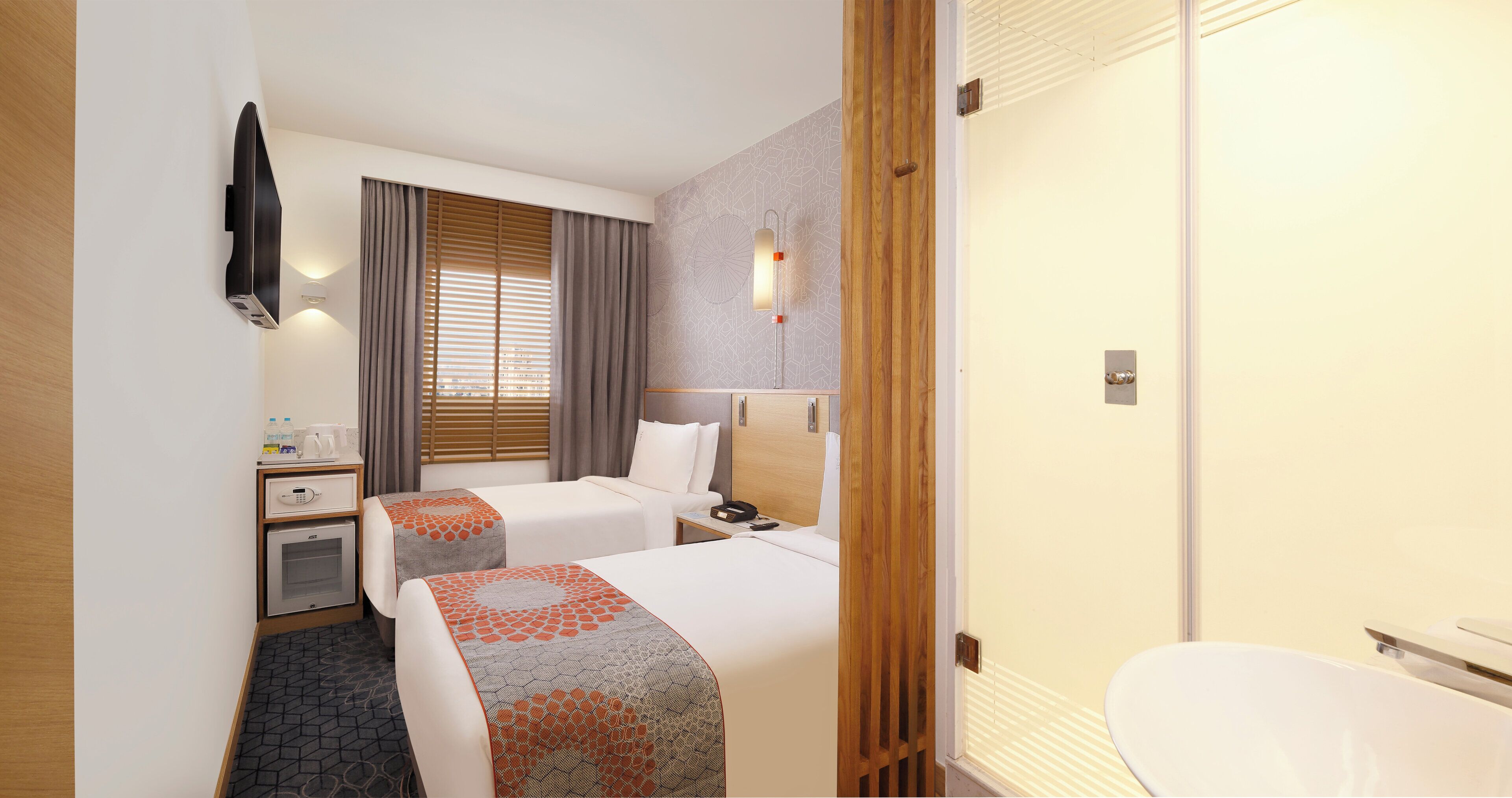 A hotel room with two beds, a desk, a minibar, and a bathroom with a toilet and sink.