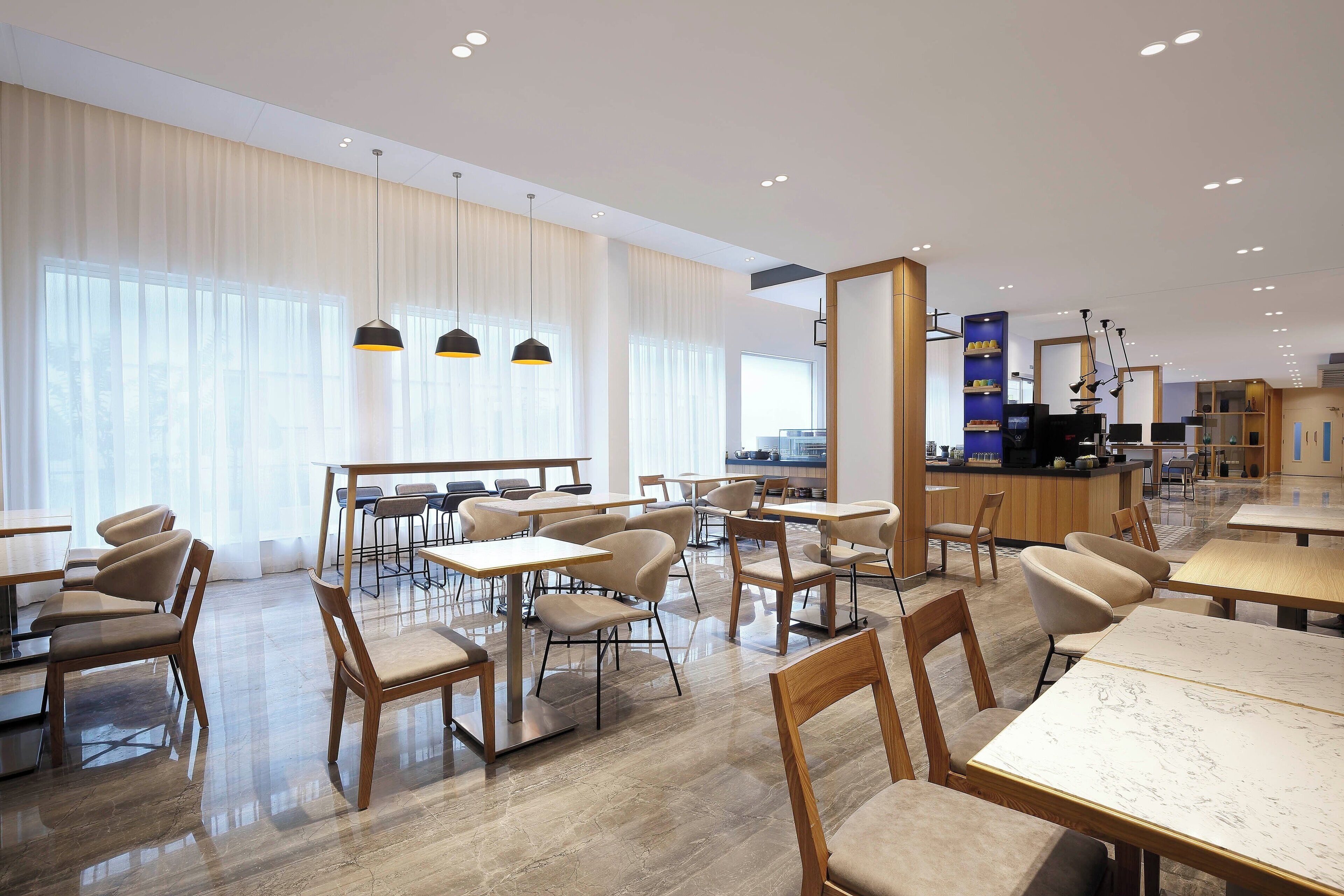 Amplia zona de comedor de hotel con varias mesas y sillas, una cocina al fondo y grandes ventanales con cortinas translúcidas.