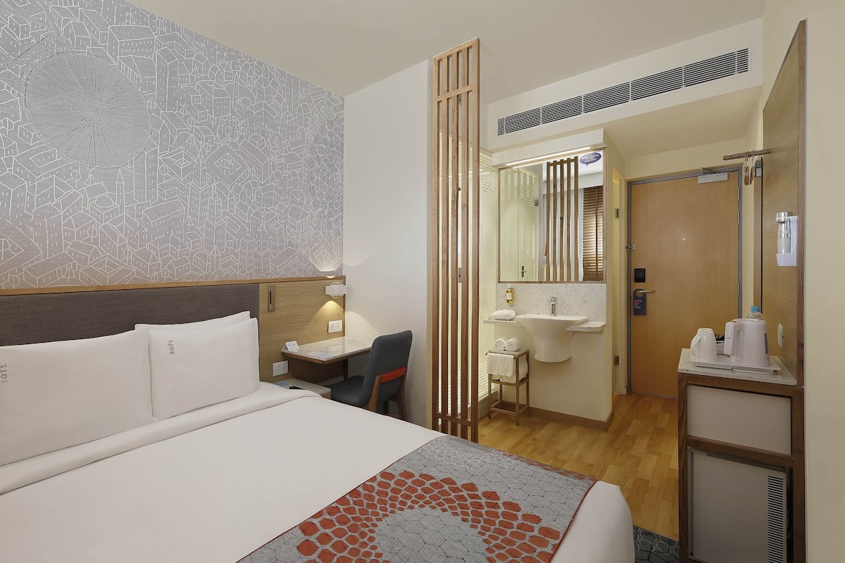 A hotel room with a bed, bedside table, lamp, and a bathroom with a sink, mirror, and towel rack.
