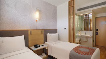 Ein Hotelzimmer mit zwei Betten, einem Nachttisch mit Lampe, einem Spiegel, einem kleinen Badezimmer mit Waschbecken und einer Wand mit geometrischem Muster.