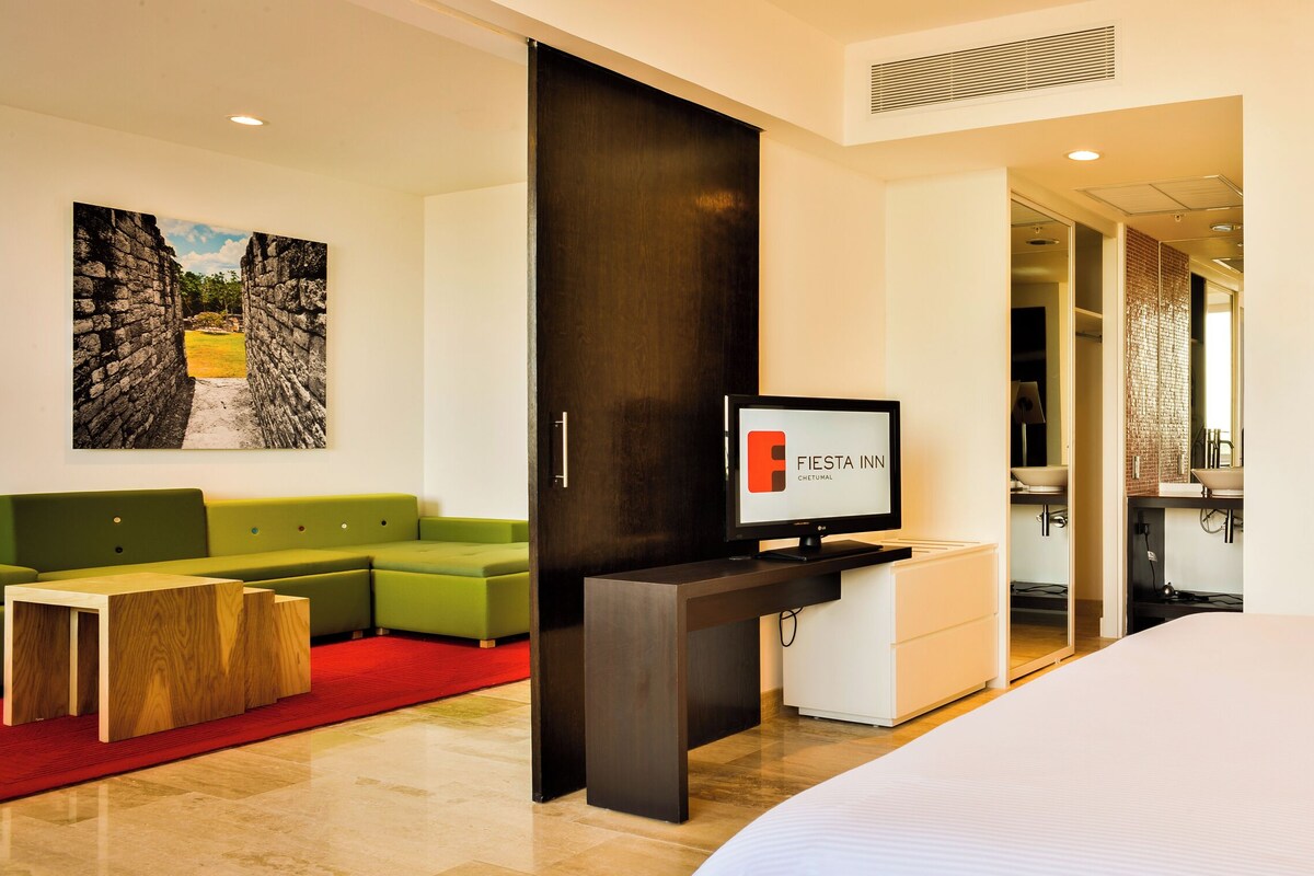 Een hotelkamer met een bed, een groene bank, een houten salontafel, een televisie en kunstwerken aan de muur.