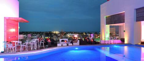 Zona de piscina en la azotea con sillones y mesas, rodeada de arquitectura moderna y vistas a la ciudad.