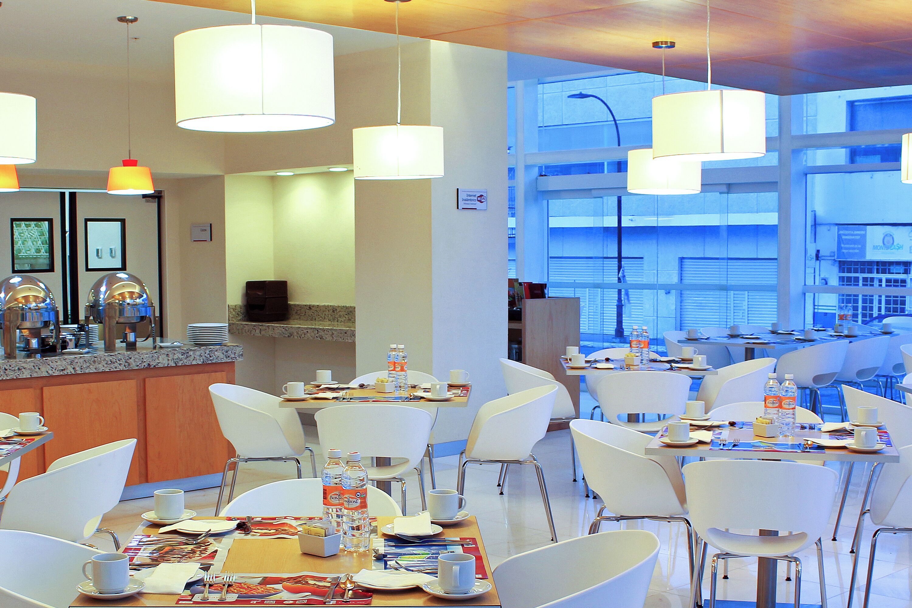 A modern hotel dining area with white chairs, tables set with cups, plates, and cutlery, and a counter with a coffee machine and water bottles.