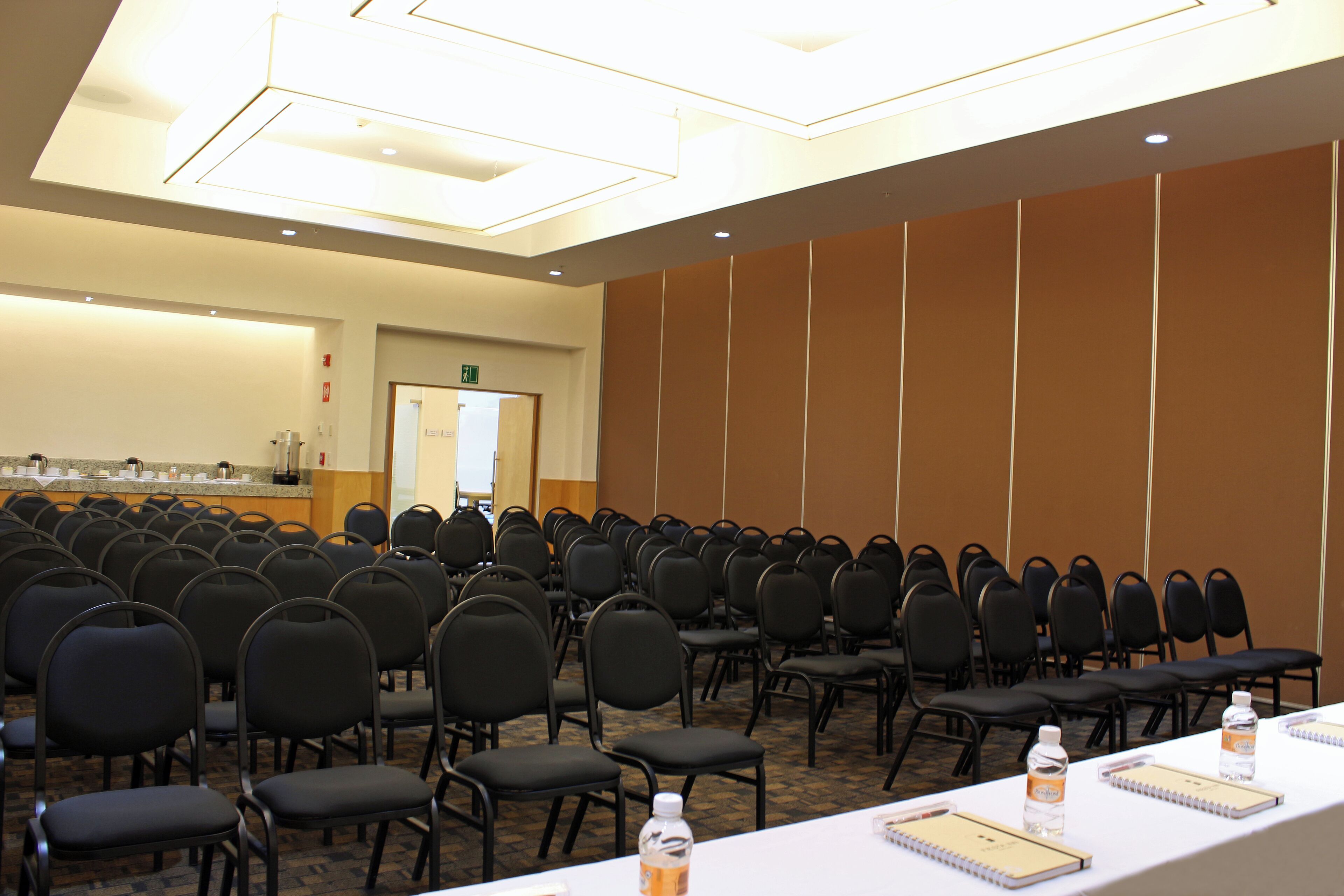 A conference room with rows of chairs, a long table with water bottles and notebooks, and a whiteboard in the background.