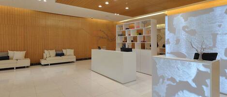 Un área de recepción moderna con techo de madera, mostrador blanco y zona de estar con sofás.