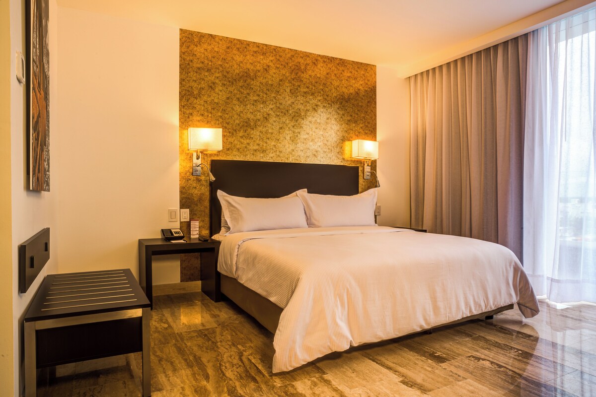 Een hotelkamer met een bed, twee bedlampen, een nachtkastje, een bureau en een groot raam met gordijnen.