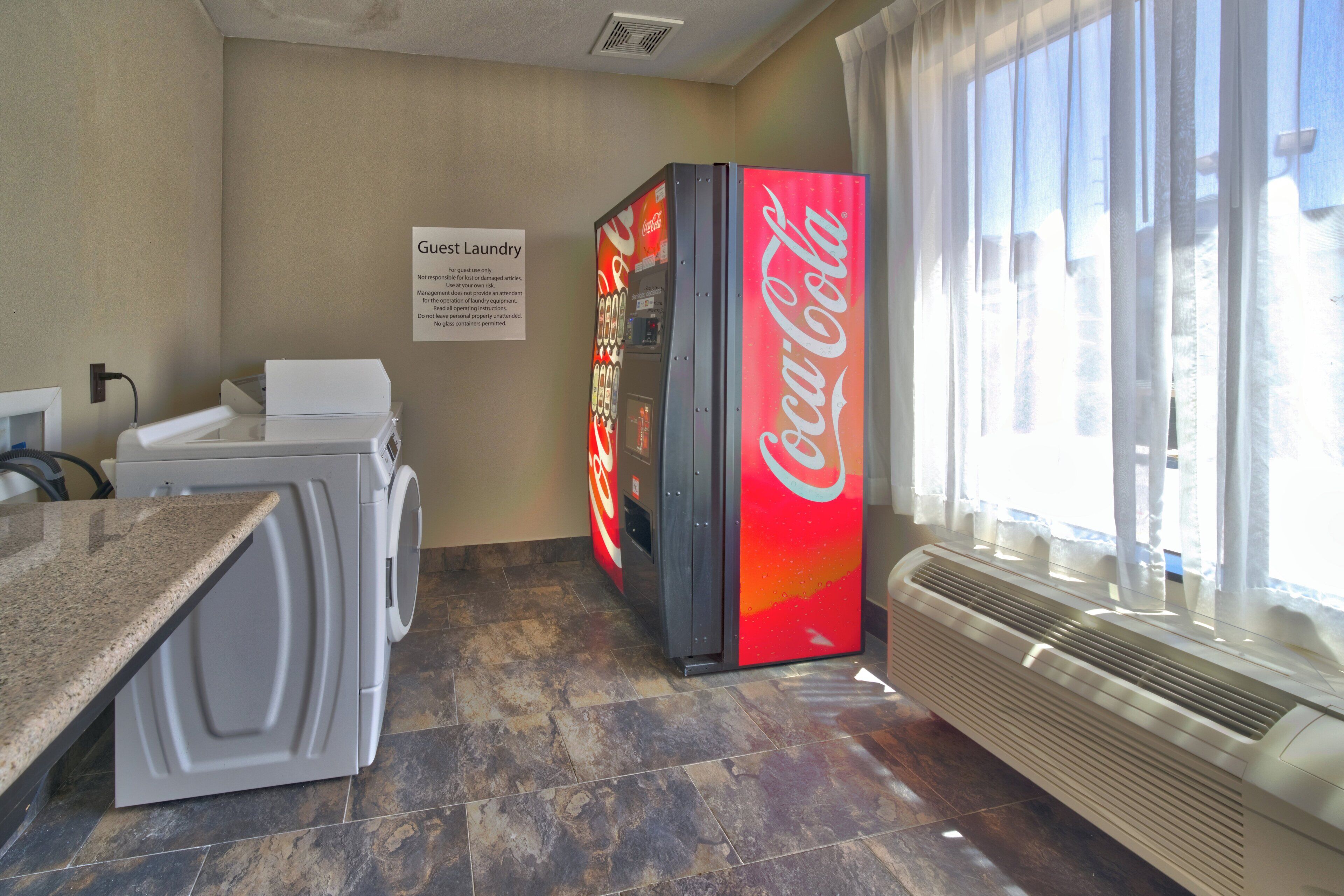 Un espace dédié à la lessive, avec un distributeur automatique de Coca-Cola, un panneau indiquant l’emplacement de la laverie pour les clients et une fenêtre avec des rideaux.