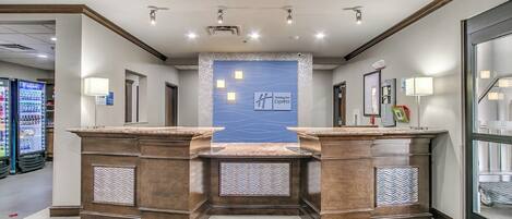 A hotel lobby with a reception desk, a blue accent wall, and a glass door leading to a parking area.