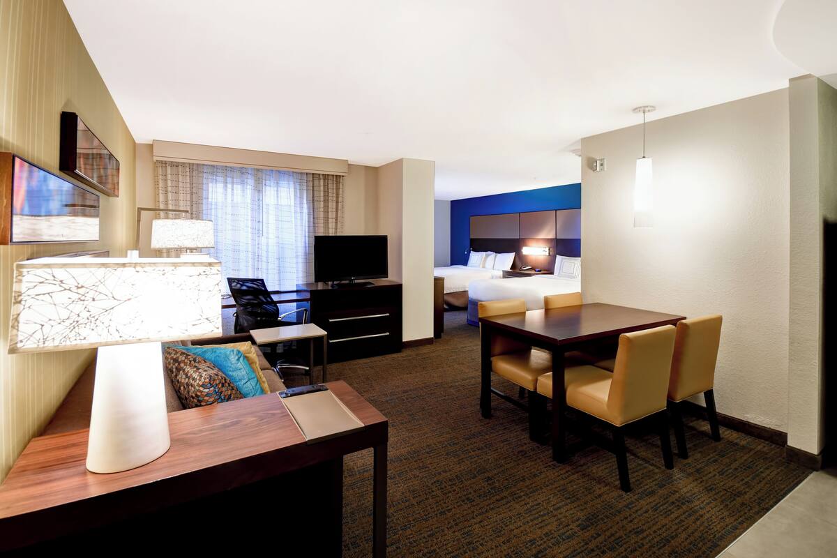 Habitación de hotel con cama, escritorio con lámpara, televisor y zona de comedor con sillas.