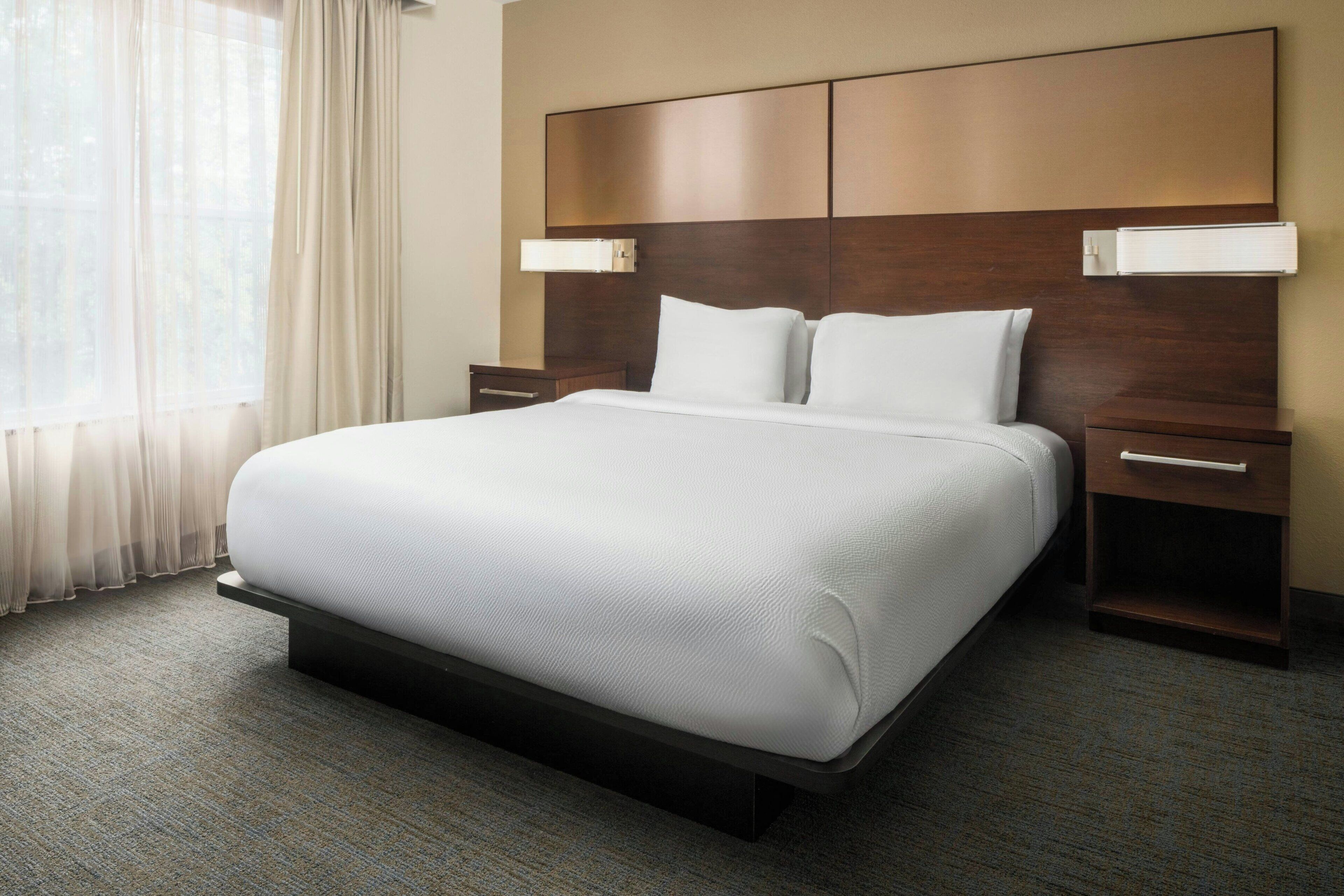 A hotel room with a large bed, two bedside tables, a headboard with integrated lighting, and a view of the outdoors through sheer curtains.