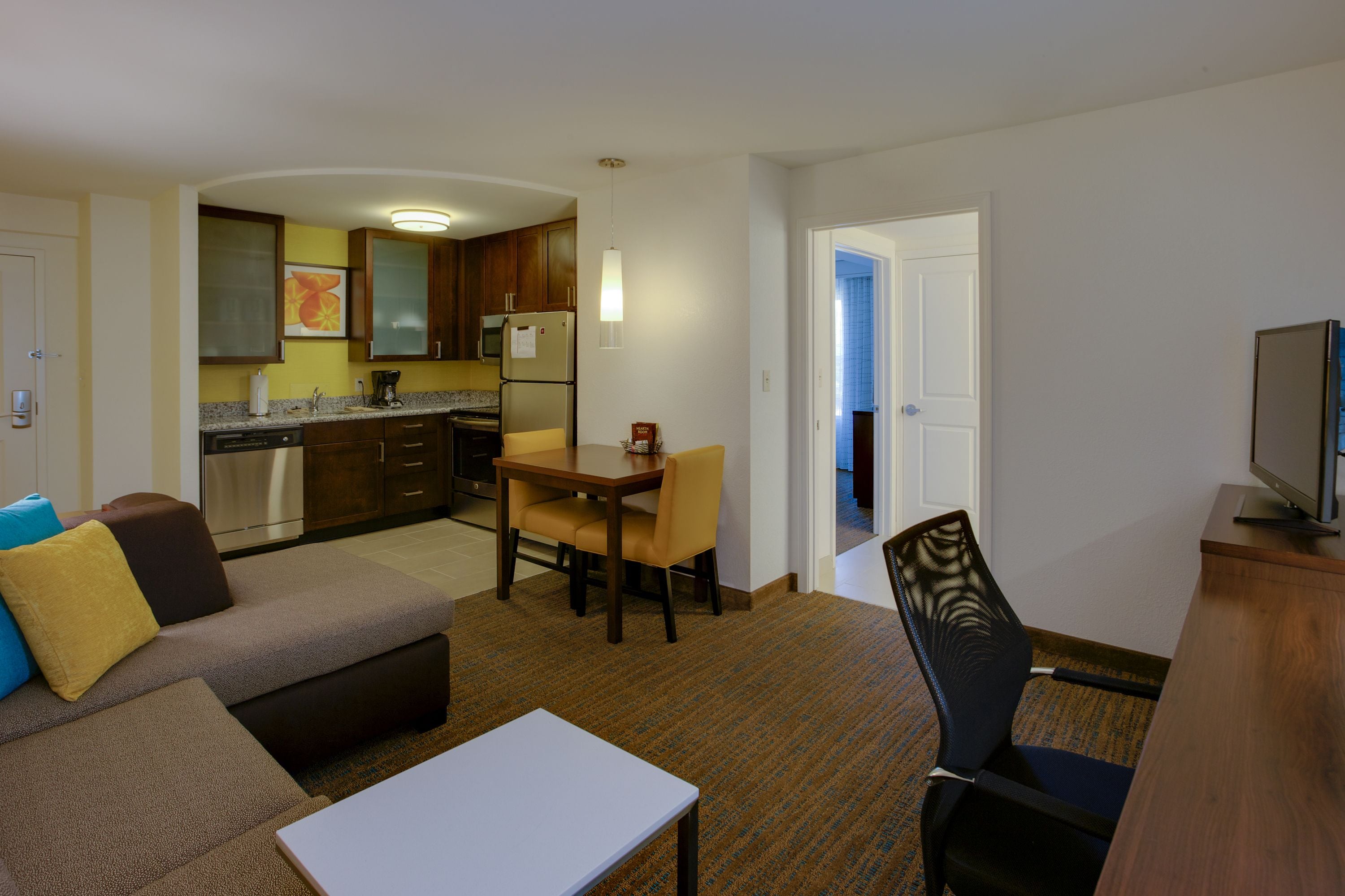 A compact hotel room with a kitchenette, a dining area with a table and chairs, a sofa, and a TV.