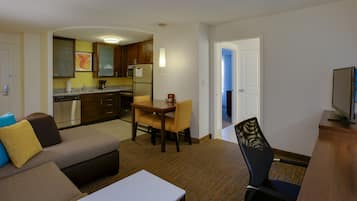 A compact hotel room with a kitchenette, a dining area with a table and chairs, a sofa, and a TV.