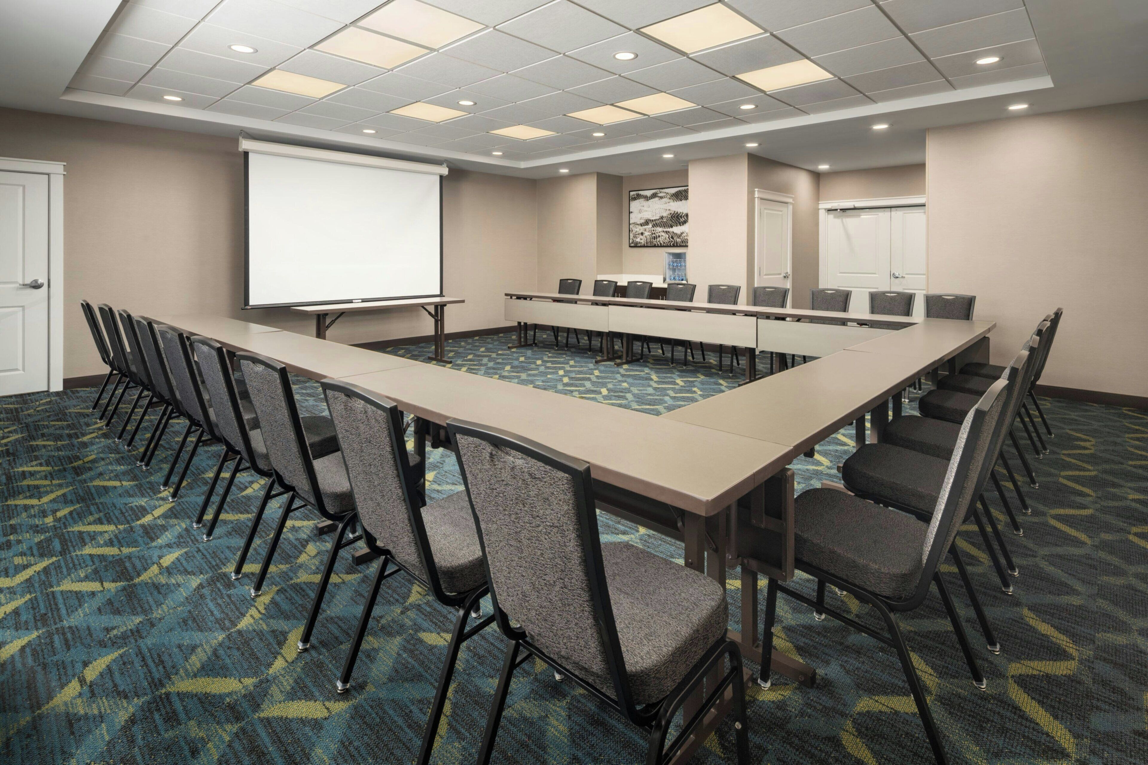 A conference room with a long table, chairs, a projector screen, and a carpeted floor.