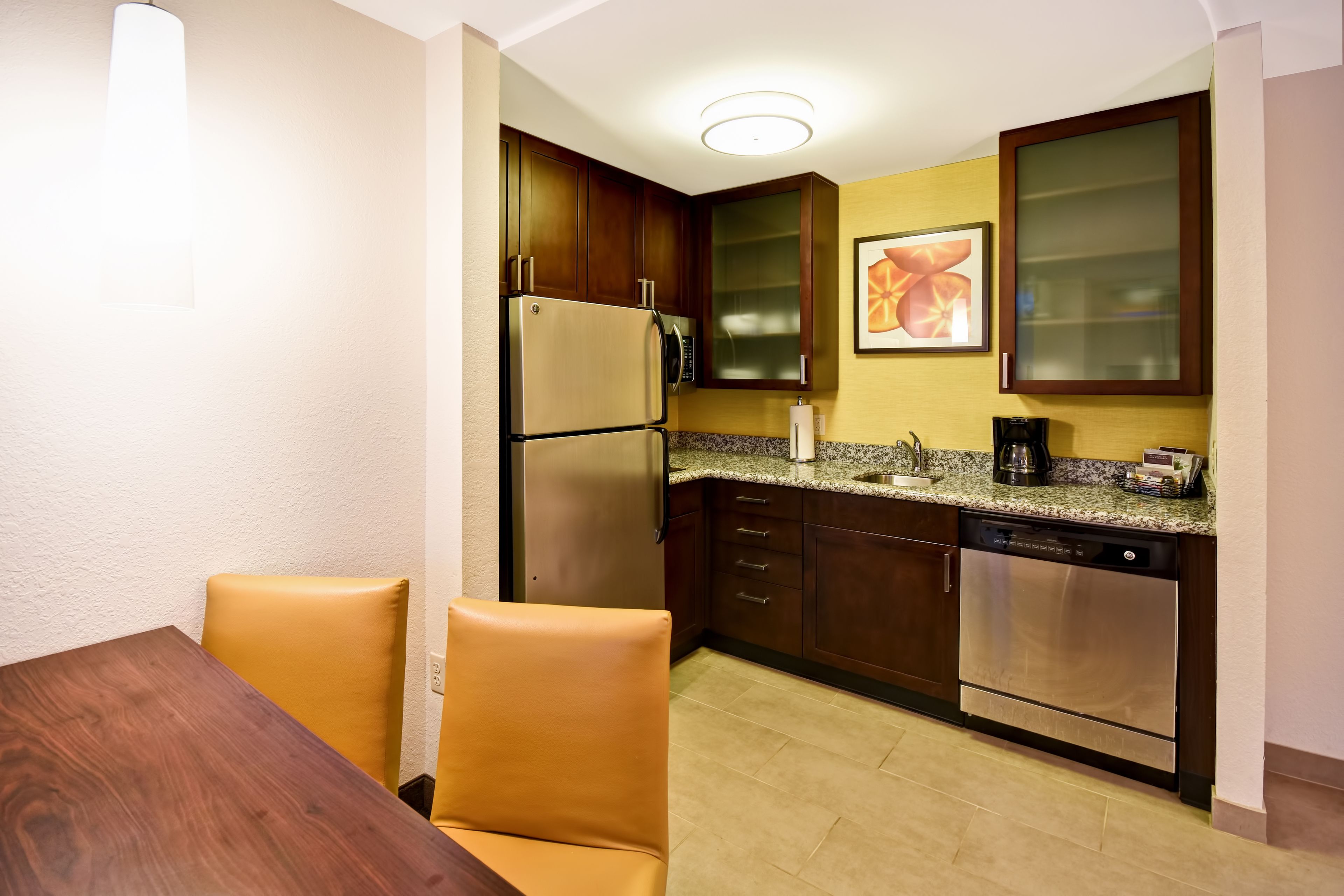 A compact kitchen with wooden cabinets, a stainless steel refrigerator, a dishwasher, and a granite countertop.
