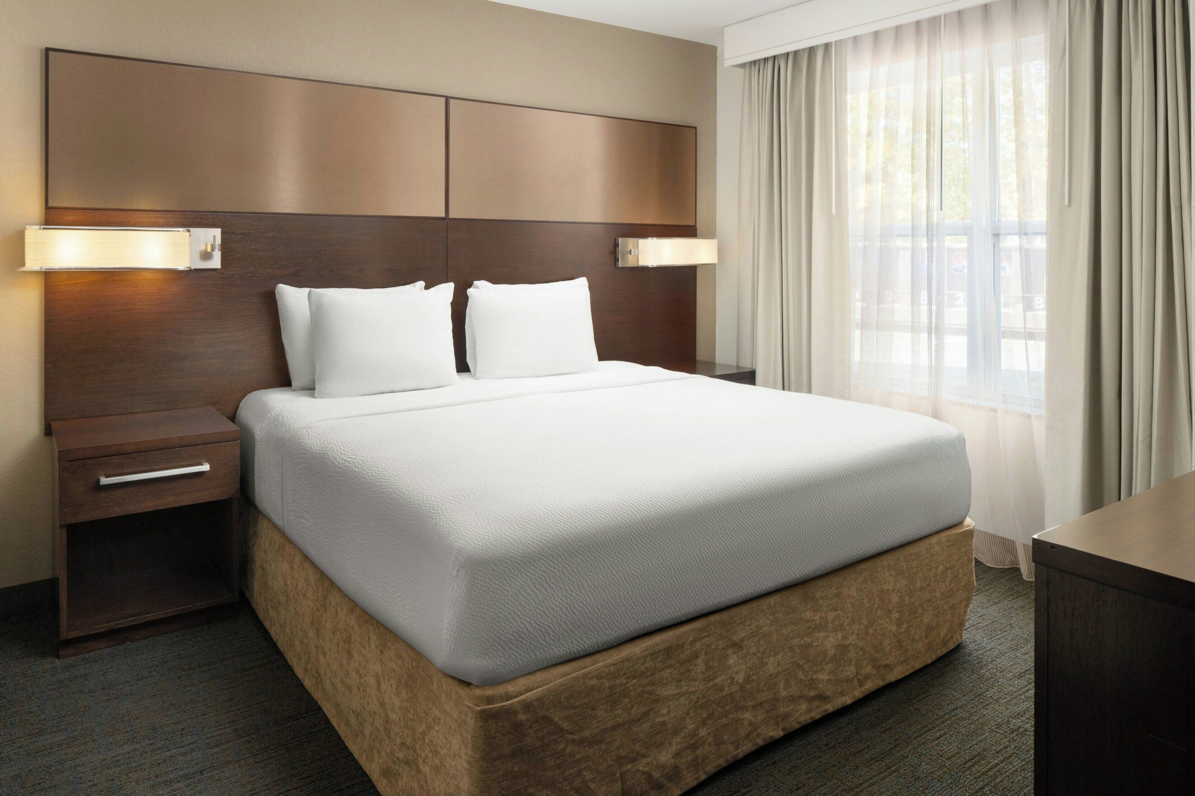 A hotel room with a large bed, wooden headboard, two bedside tables with lamps, and a window with curtains.