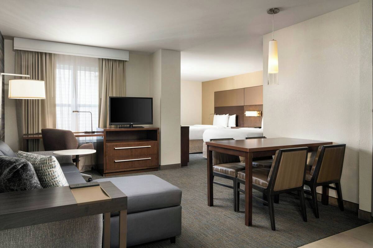 Habitación de hotel con sofá, una mesa de comedor con sillas, televisor, escritorio con lámpara y cama.