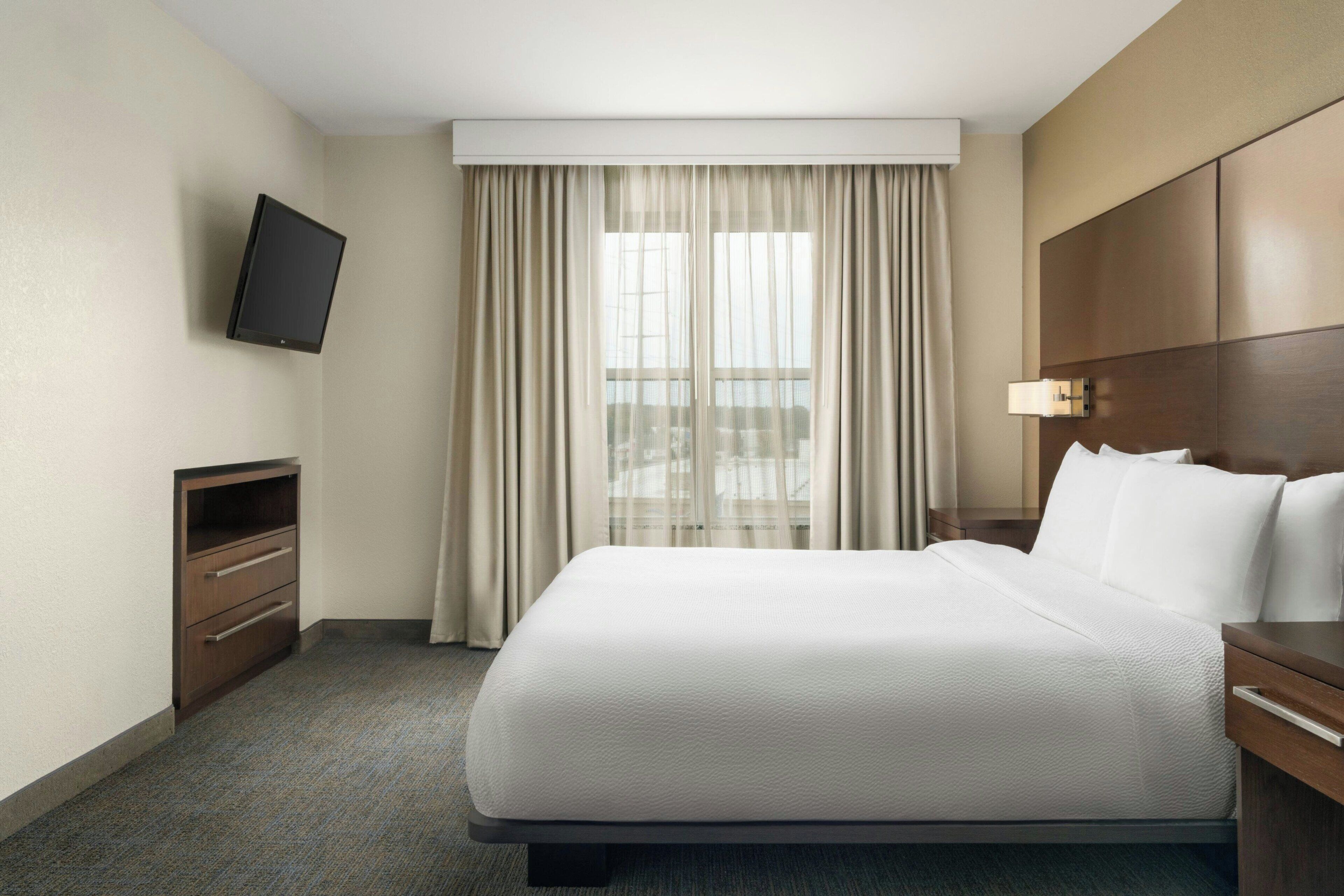 A hotel room with a large bed, a TV mounted on the wall, a desk with a lamp, and a window with curtains.