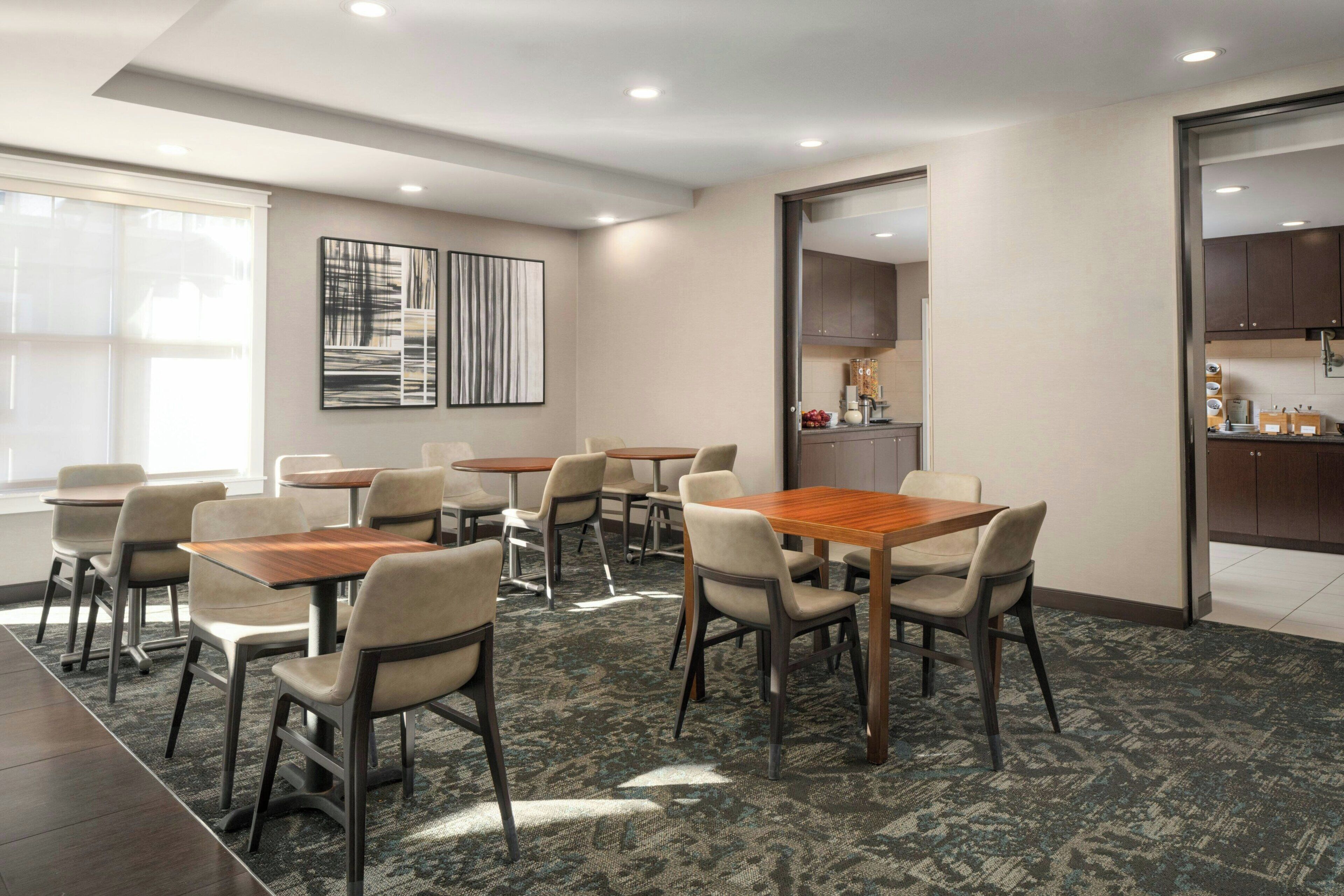A conference room with multiple tables and chairs, a large window, and a kitchen area visible through an open door.