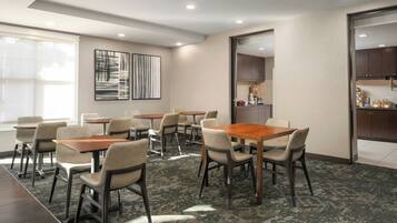 A conference room with multiple tables and chairs, a large window, and a kitchen area visible through an open door.