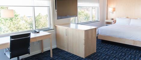 Habitación de hotel con cama, escritorio con silla, televisor montado en la pared y ventana con vista a árboles.