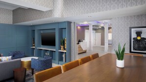 A modern living room with a large wooden dining table, blue and grey furniture, and a flat-screen TV mounted on the wall.