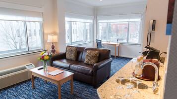 Habitación de hotel con un sofá de cuero marrón, una mesita con un florero, un escritorio con una silla y una kitchenette con un lavamanos y encimera.