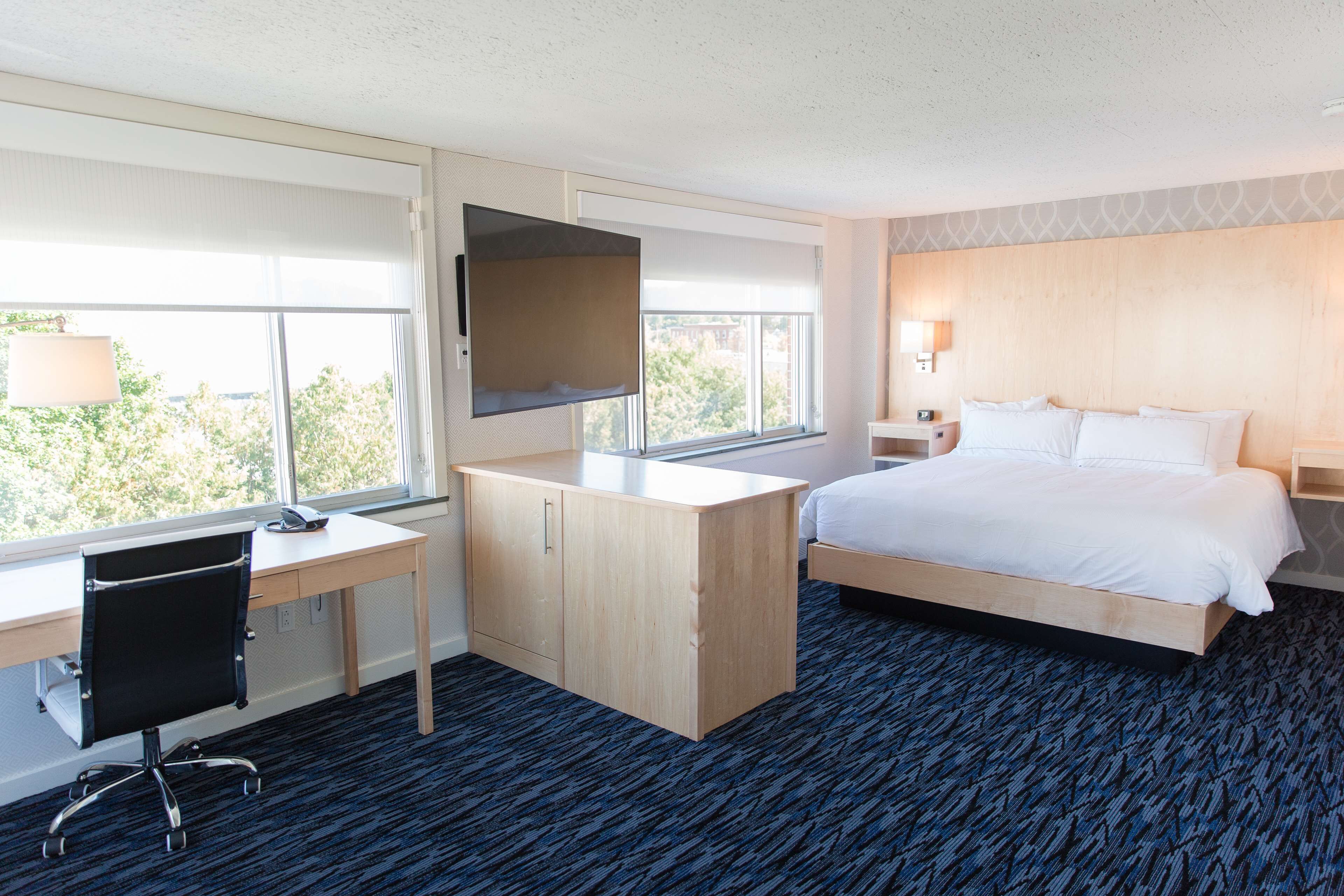 A hotel room with a bed, desk, chair, TV, and large windows.