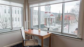 A room with a wooden desk, a white chair, and large windows offering a view of buildings and a clock tower.