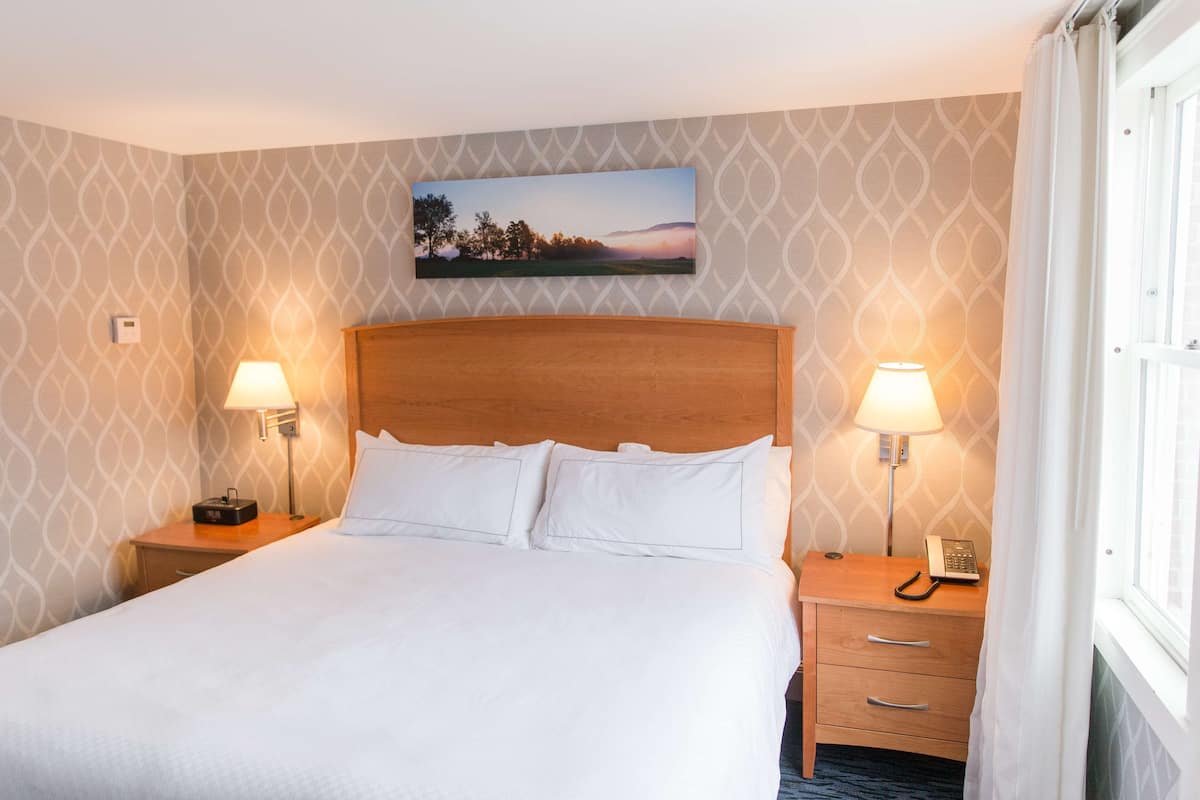 Ein Hotelzimmer mit einem großen Bett, zwei Nachttischen mit Lampen, einem Holzkopfstück, einem Bild an der Wand und einem Fenster mit Vorhängen.