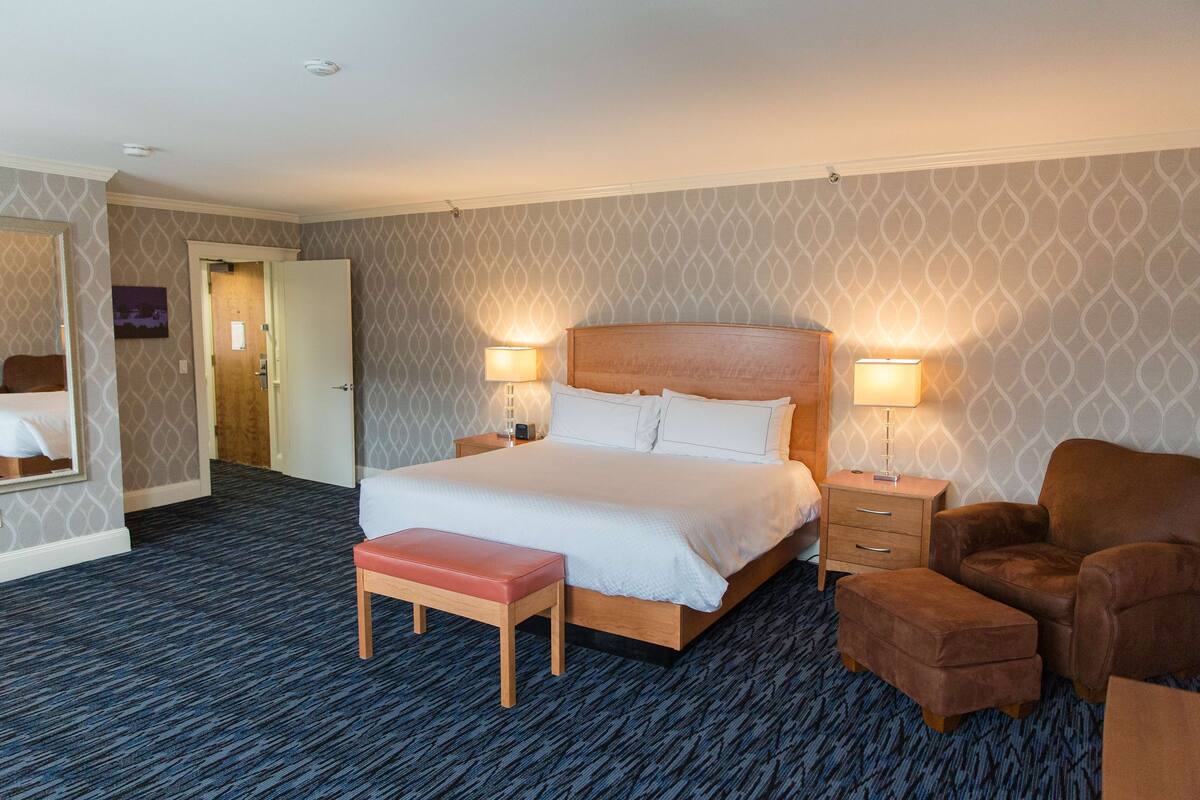 A hotel room with a large bed, two bedside tables with lamps, a wooden bench, and a brown armchair.