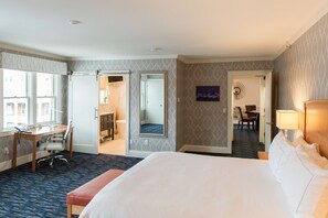 A hotel room with a large bed, a desk with a chair, a mirror, and a dining area visible through an open door.
