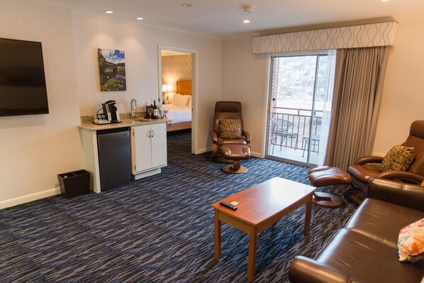 Habitación de hotel con kitchenette, sala de estar con sofá y sillón, una pequeña mesa y balcón con vistas.