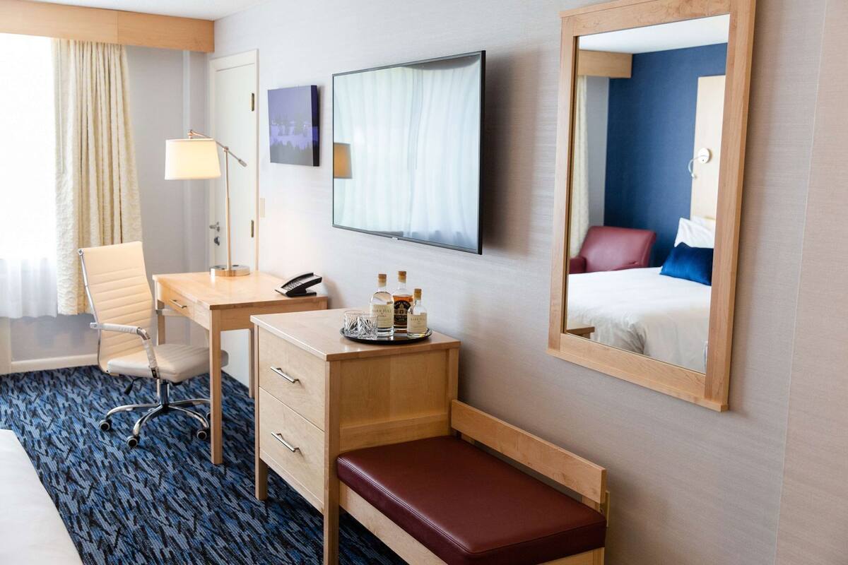 A hotel room with a desk, chair, bedside table, lamp, and a mirror reflecting the bed and room.