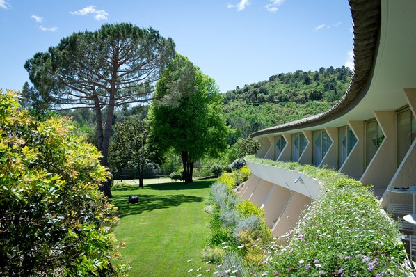 A hotel with a curved roof, a garden with various plants and a large tree, and a view of a hilly landscape. - La Vague de Saint Paul (Vence)