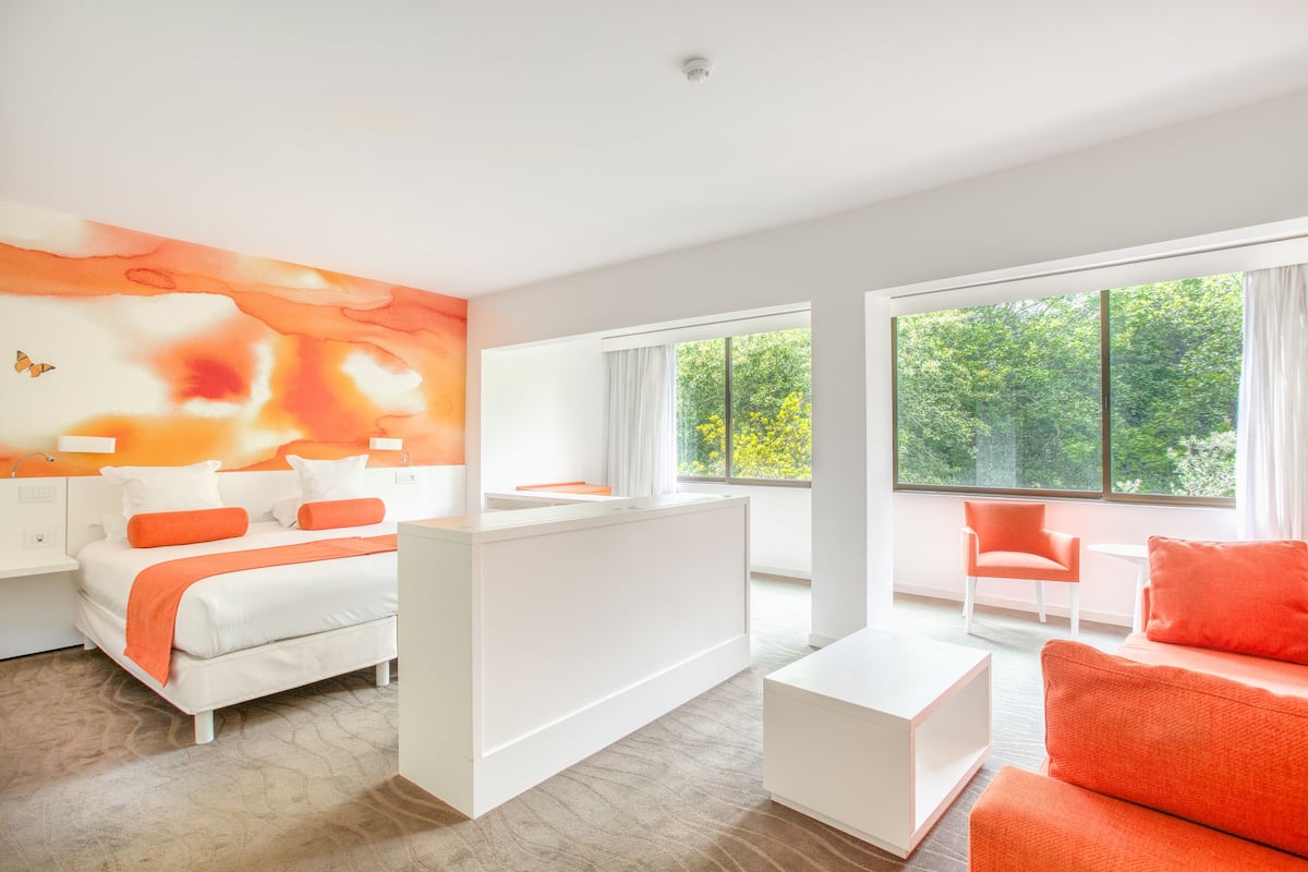 Une chambre d’hôtel moderne avec un grand lit, un bureau blanc, un canapé orange vif et de grandes fenêtres donnant sur un paysage verdoyant.