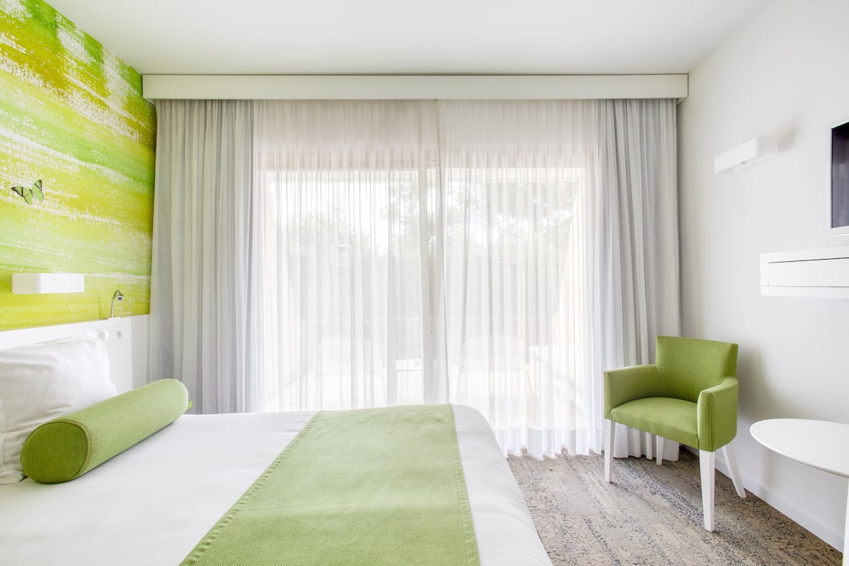 Une chambre d’hôtel avec un grand lit, un fauteuil vert, une table blanche et une fenêtre ornée de rideaux légers.